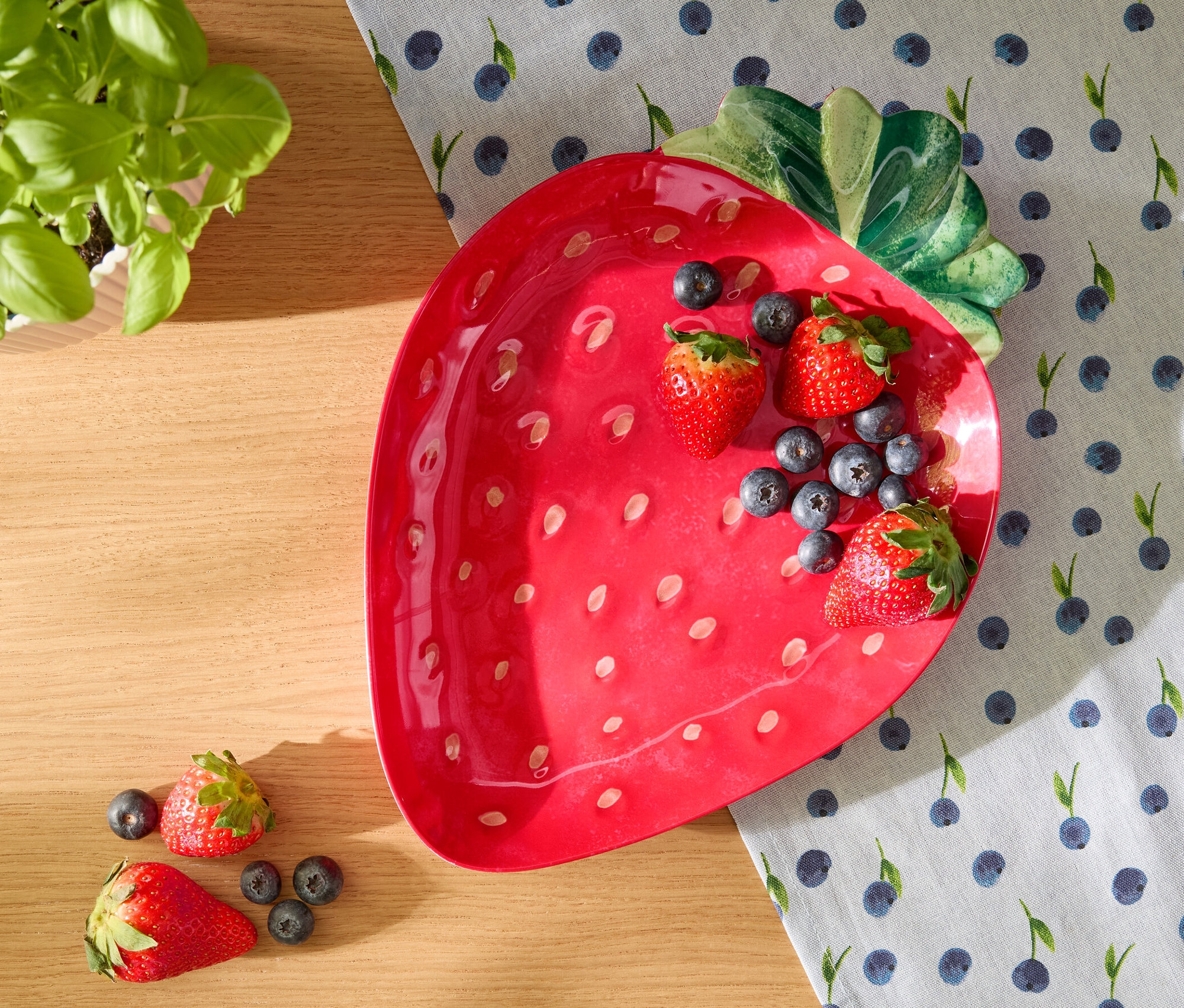 Roter Servierteller in Erdbeerform mit Erdbeeren und Blaubeeren auf einem Tischläufer mit Blaubeer-Muster.