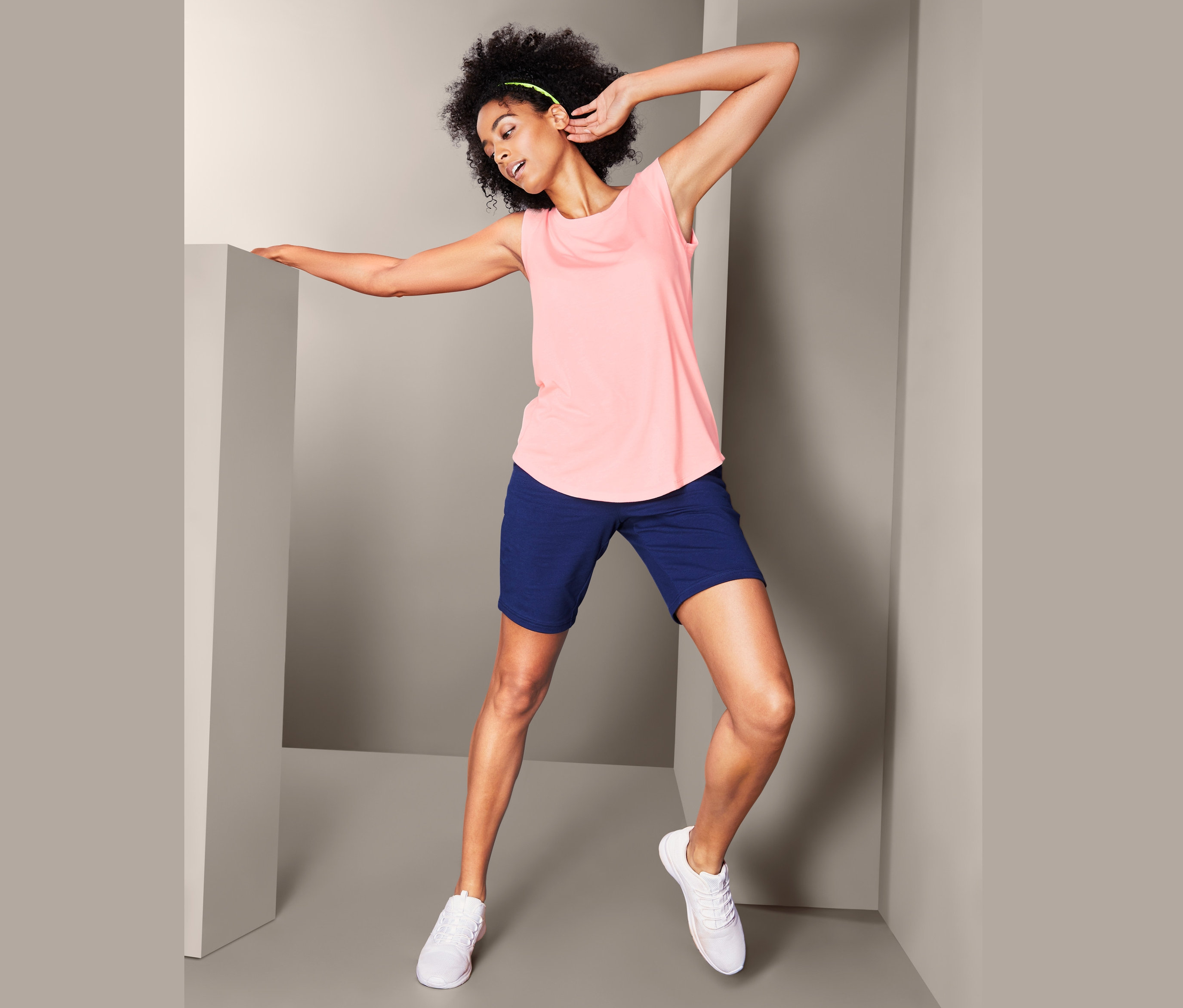 Frau posiert in einem rosafarbenen Top und blauen Shorts. Sie trägt weiße Turnschuhe. 2 Tops