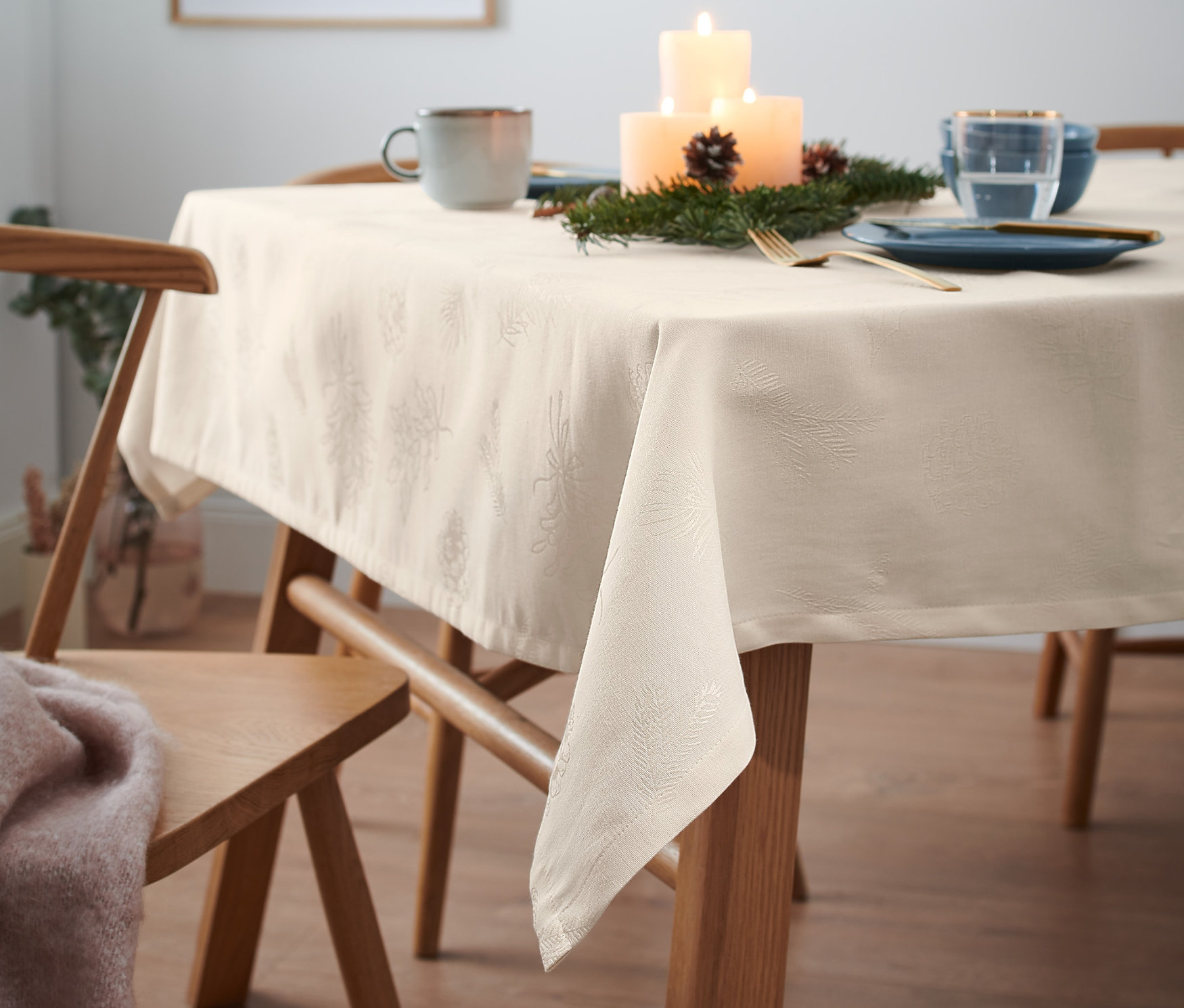 Gedeckter Tisch mit cremefarbener Jacquard-Tischdecke, Tellern, Tasse, Kerzen und Dekorationen.