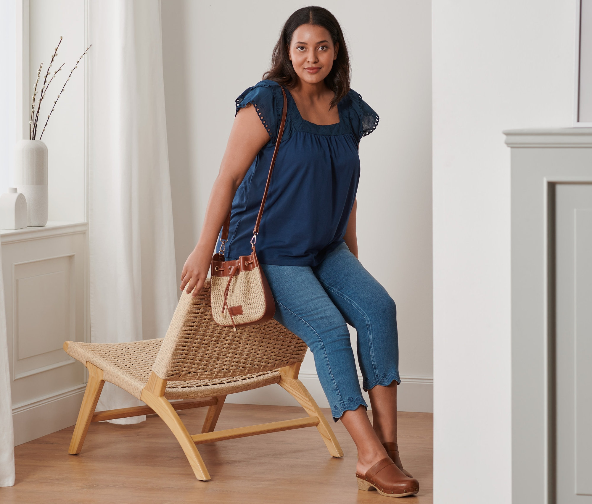 Eine Frau sitzt auf einem Korbstuhl und trägt ein dunkelblaues Shirt mit Stickerei, 7/8-Treggings mit Lochstickerei und Leder-Clogs. Sie hat eine Beuteltasche dabei.
