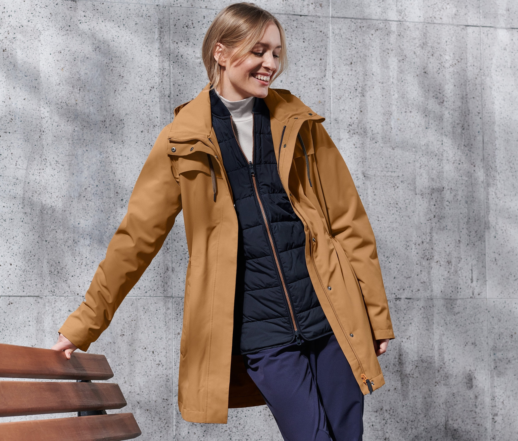 Eine lächelnde Frau lehnt an einer Bank und trägt einen cognacfarbenen 3-in-1-Outdoormantel.