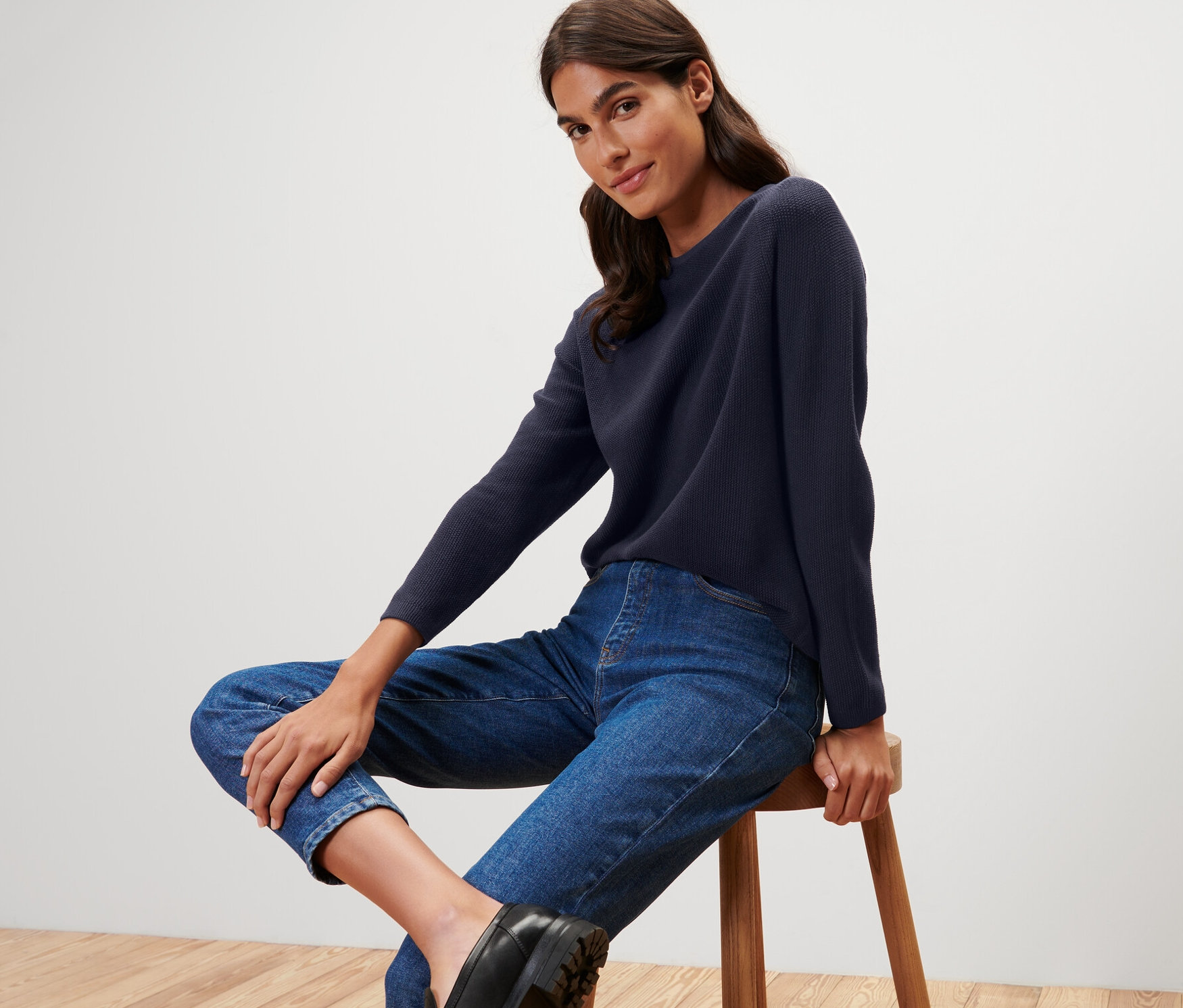 Eine Frau sitzt auf einem Hocker und trägt einen dunkelblauen Strickpullover.