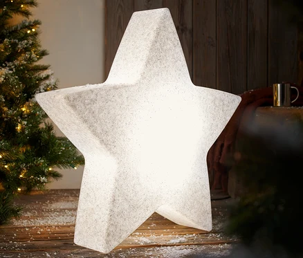 Eine leuchtende Smart-Outdoorleuchte »Stern«, L, steht auf einem schneebedeckten Holzboden neben einem Weihnachtsbaum.