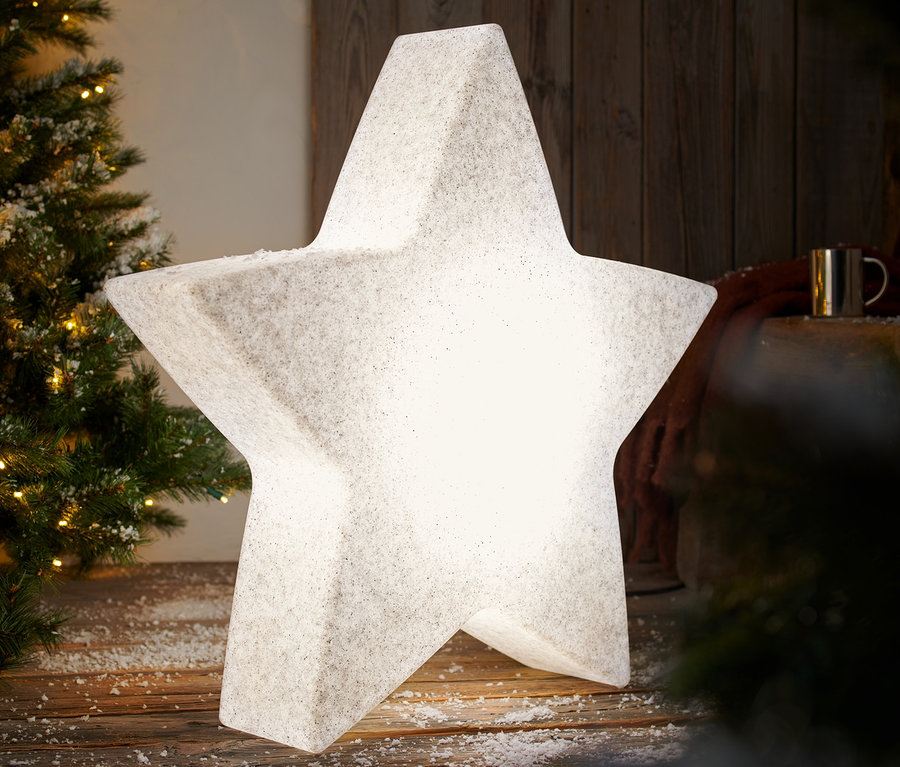 Eine leuchtende Smart-Outdoorleuchte »Stern«, L, steht auf einem schneebedeckten Holzboden neben einem Weihnachtsbaum.
