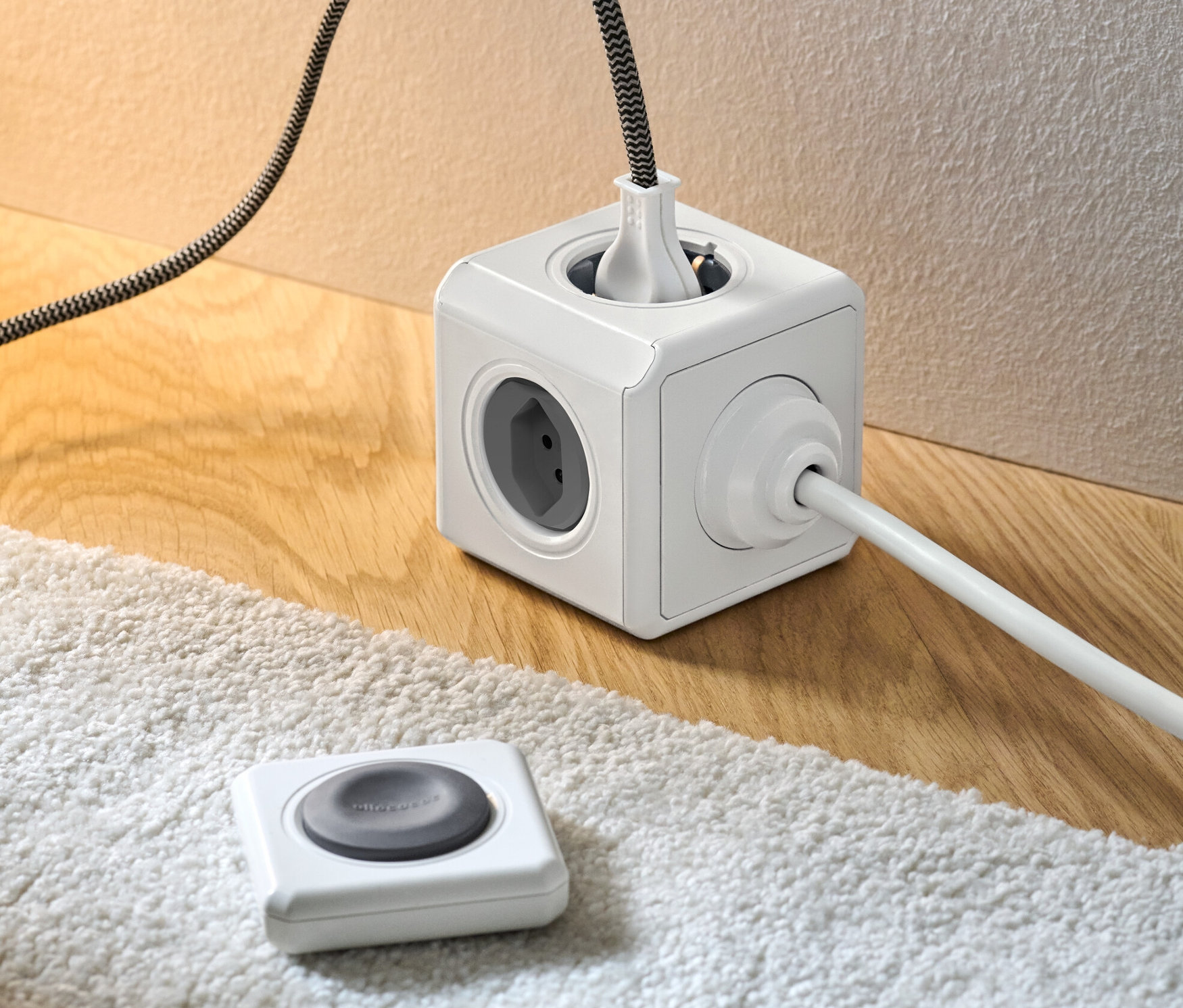Ein weißer Steckdosenwürfel mit Fernbedienung und eingesteckten Kabeln auf einem Holzboden und einem weißen Teppich.