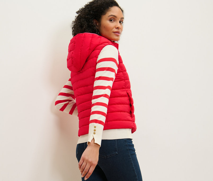 Eine Frau blickt über ihre Schulter und trägt eine Steppweste mit Kapuze, ein gestreiftes Oberteil und Jeans.