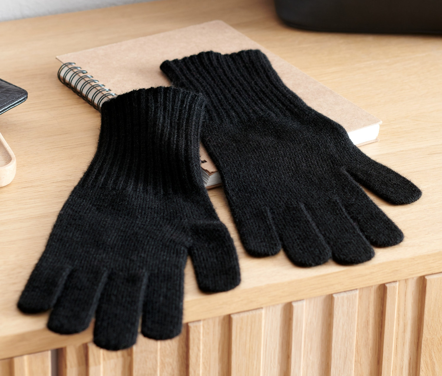 Auf einer Holzkommode liegen ein Paar schwarze Strickhandschuhe mit Wolle, ein Notizbuch und eine schwarze Handtasche.