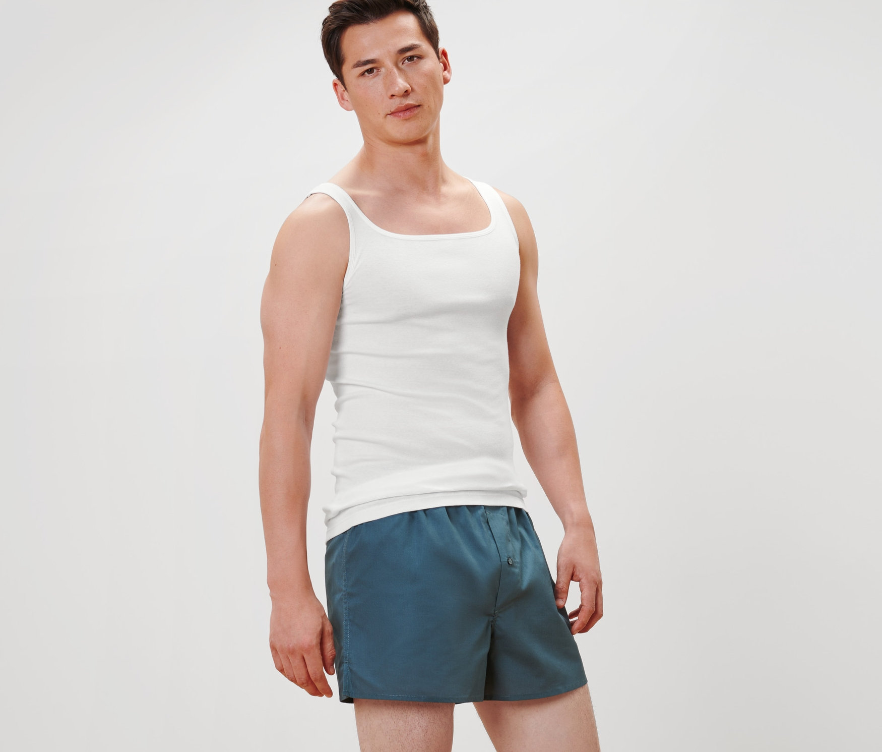 Ein Mann steht und trägt 2 Qualitäts-Feinripp-Unterhemden in Weiß und blaue Shorts.