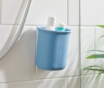 Ein blauer Bad-Organizer mit einem weißen und einem blauen Produkt darin, an weißen Fliesen neben einem Duschschlauch befestigt.