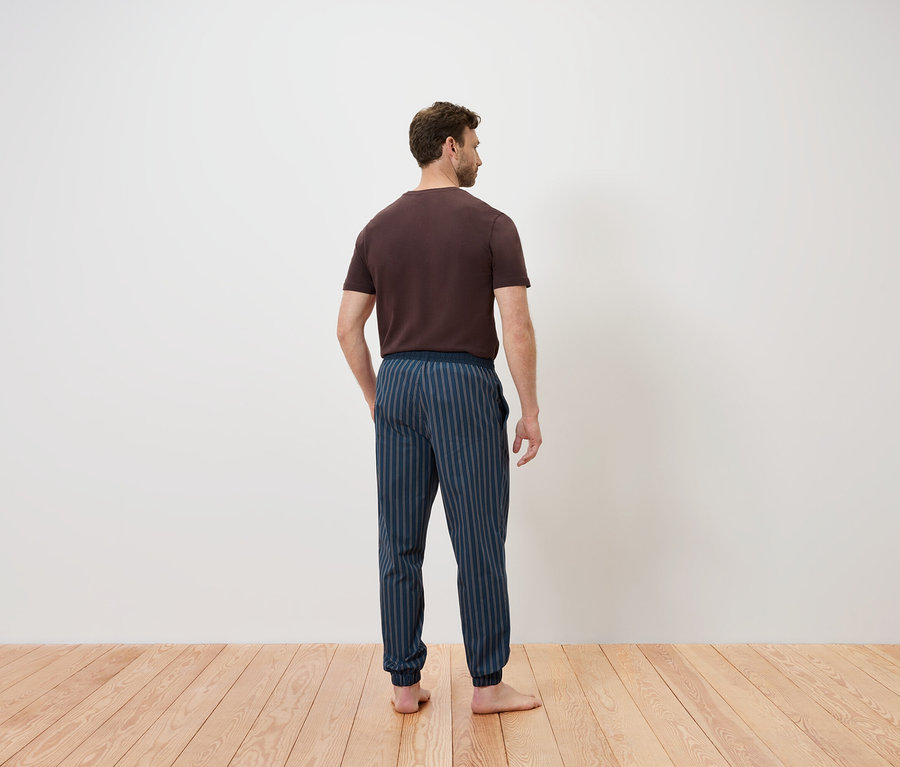 Mann mit dem Rücken zur Kamera steht auf einem Holzboden. Er trägt ein braunes T-Shirt und eine blau gestreifte Hose.