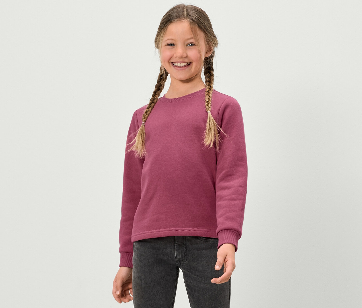 Lächelndes Mädchen mit Zöpfen in einem rosa Kinder-Sweatshirt mit angerauter Innenseite.