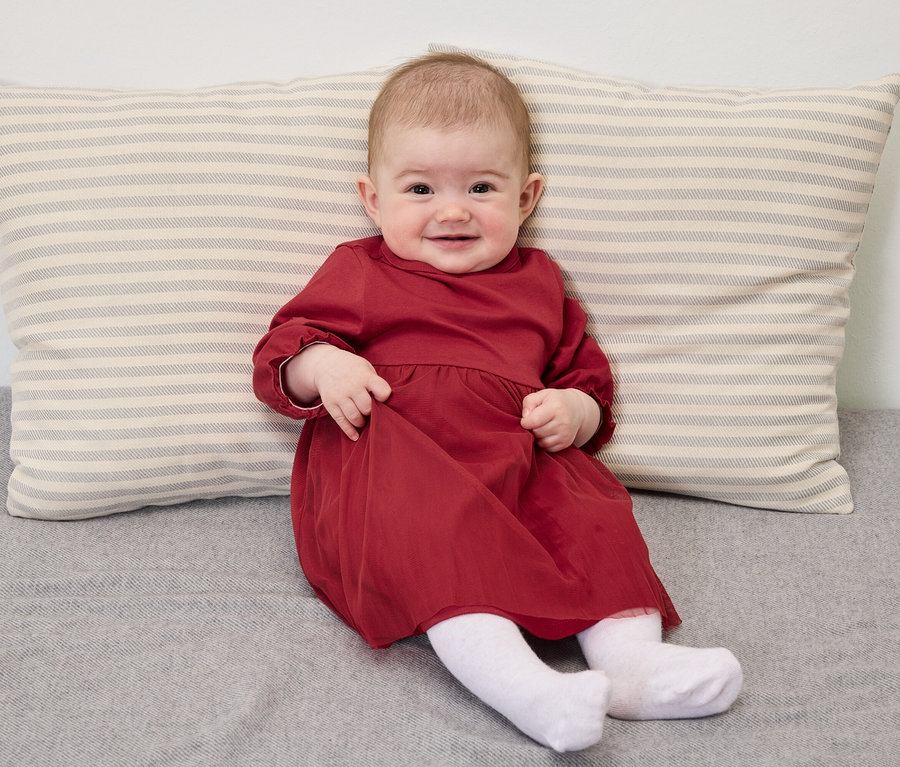 Ein lächelndes Baby in einem roten Kleid sitzt auf einer grauen Decke mit einem gestreiften Kissen.