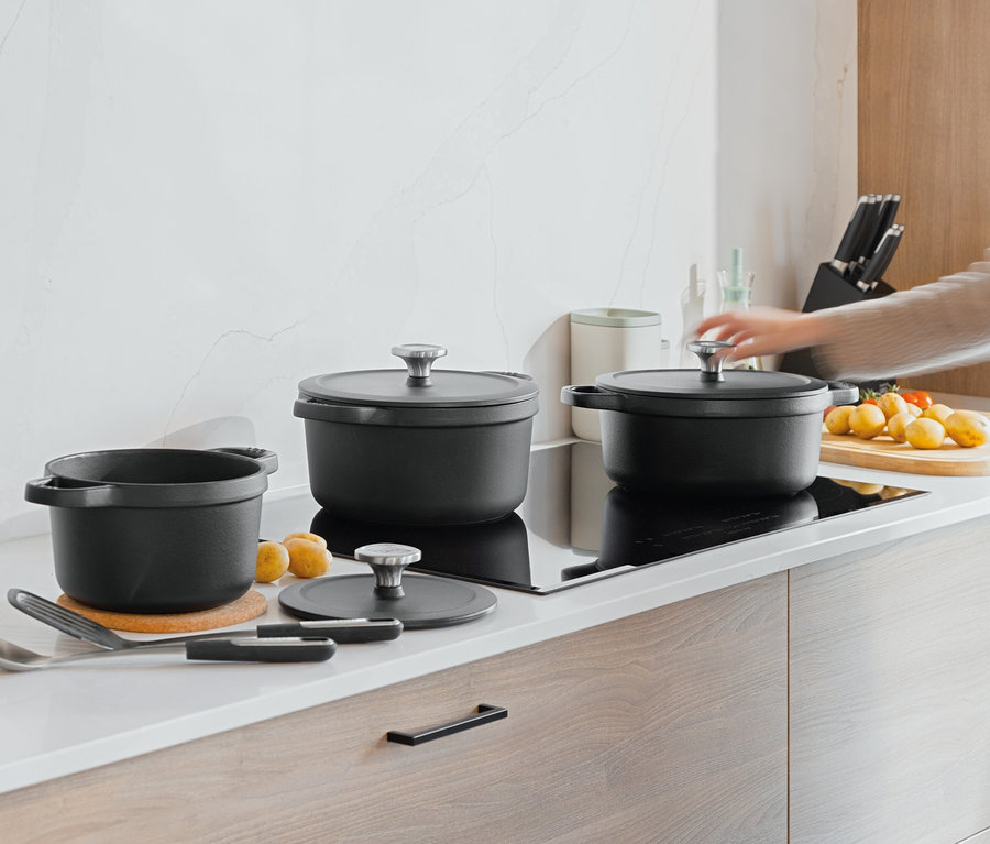 Auf einer Küchentheke stehen drei schwarze Töpfe, daneben liegen Kartoffeln und Tomaten. Eine Hand greift nach dem Deckel eines der Töpfe.