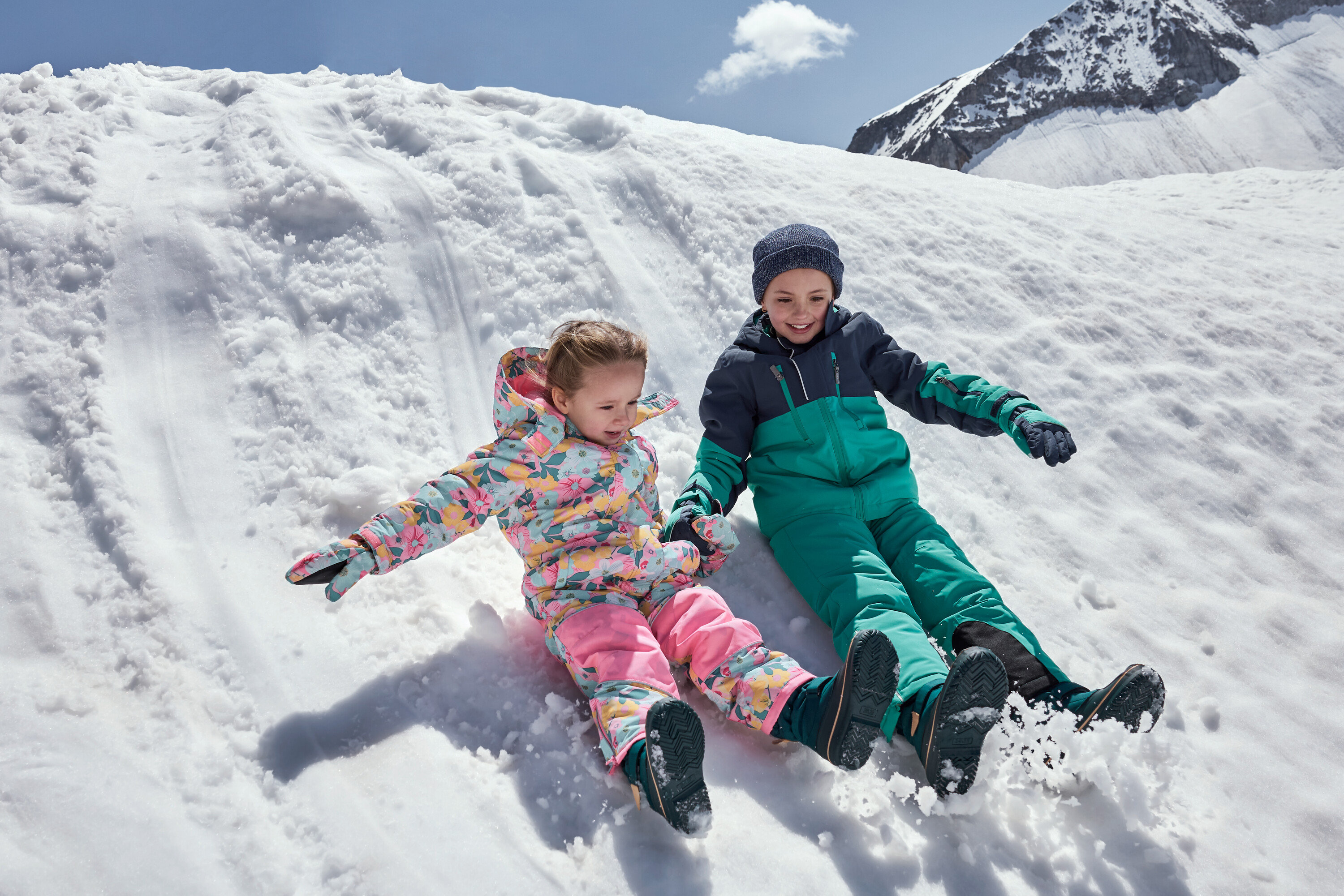 Kinder rutschen einen verschneiten Hügel hinunter, bekleidet mit farbenfrohen Winteroveralls.