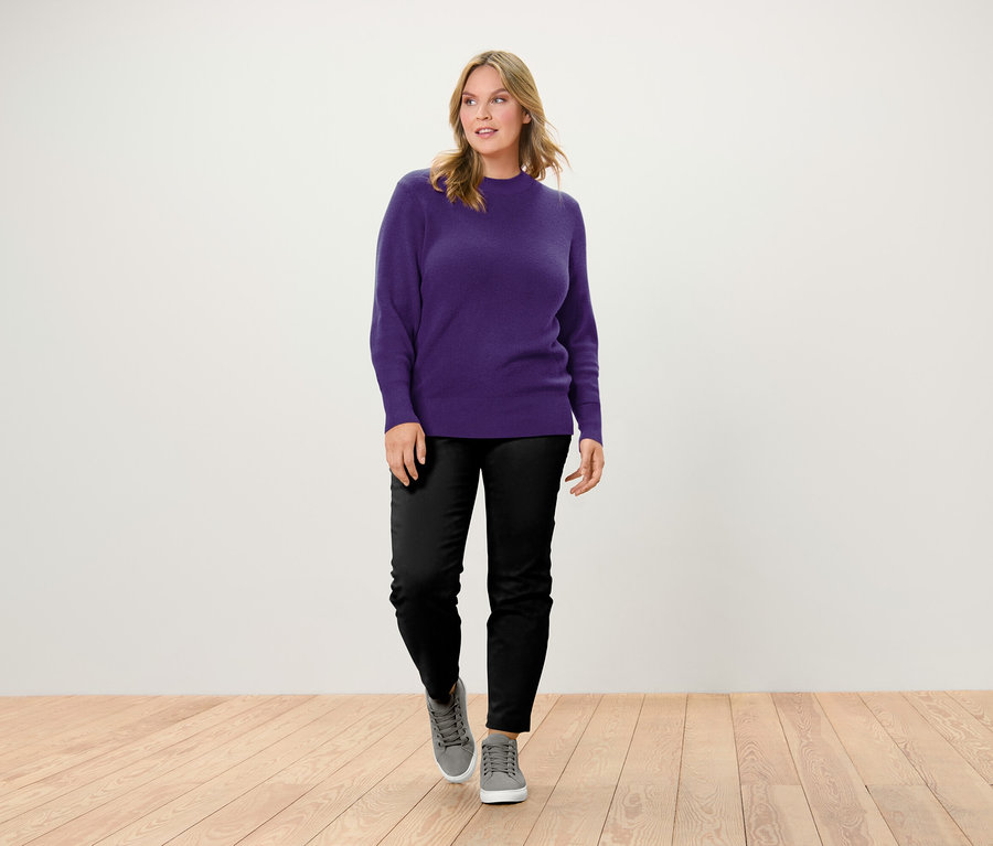 Eine Frau geht auf einem Holzboden und trägt einen lila Pullover, schwarze Hosen und graue Turnschuhe.