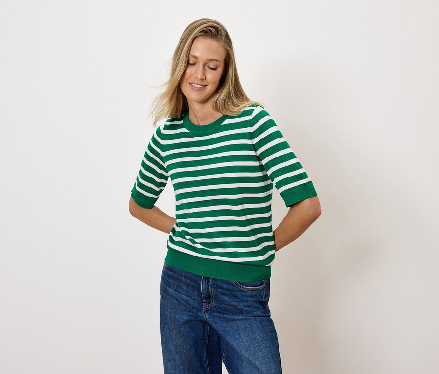 Eine blonde Frau posiert in einem grün-weiß gestreiften Oberteil und blauen Jeans.