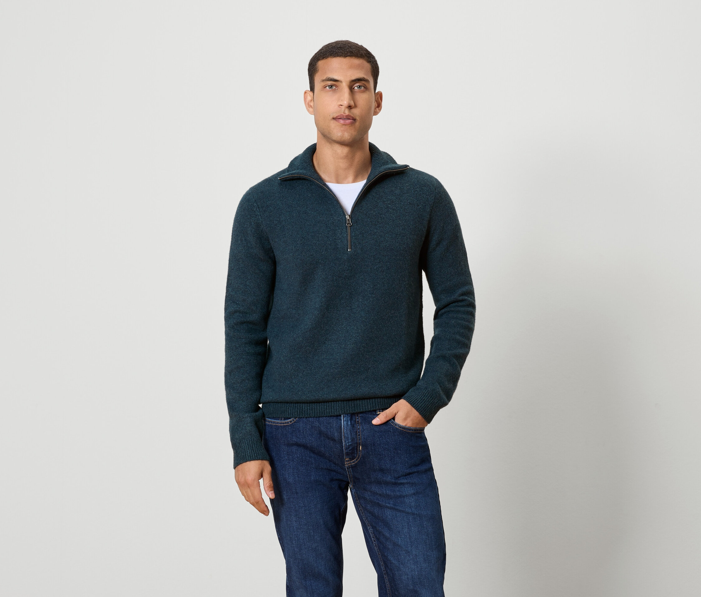 Mann posiert vor einem Hintergrund und trägt einen dunkelblauen Pullover mit Reißverschluss und Jeans.