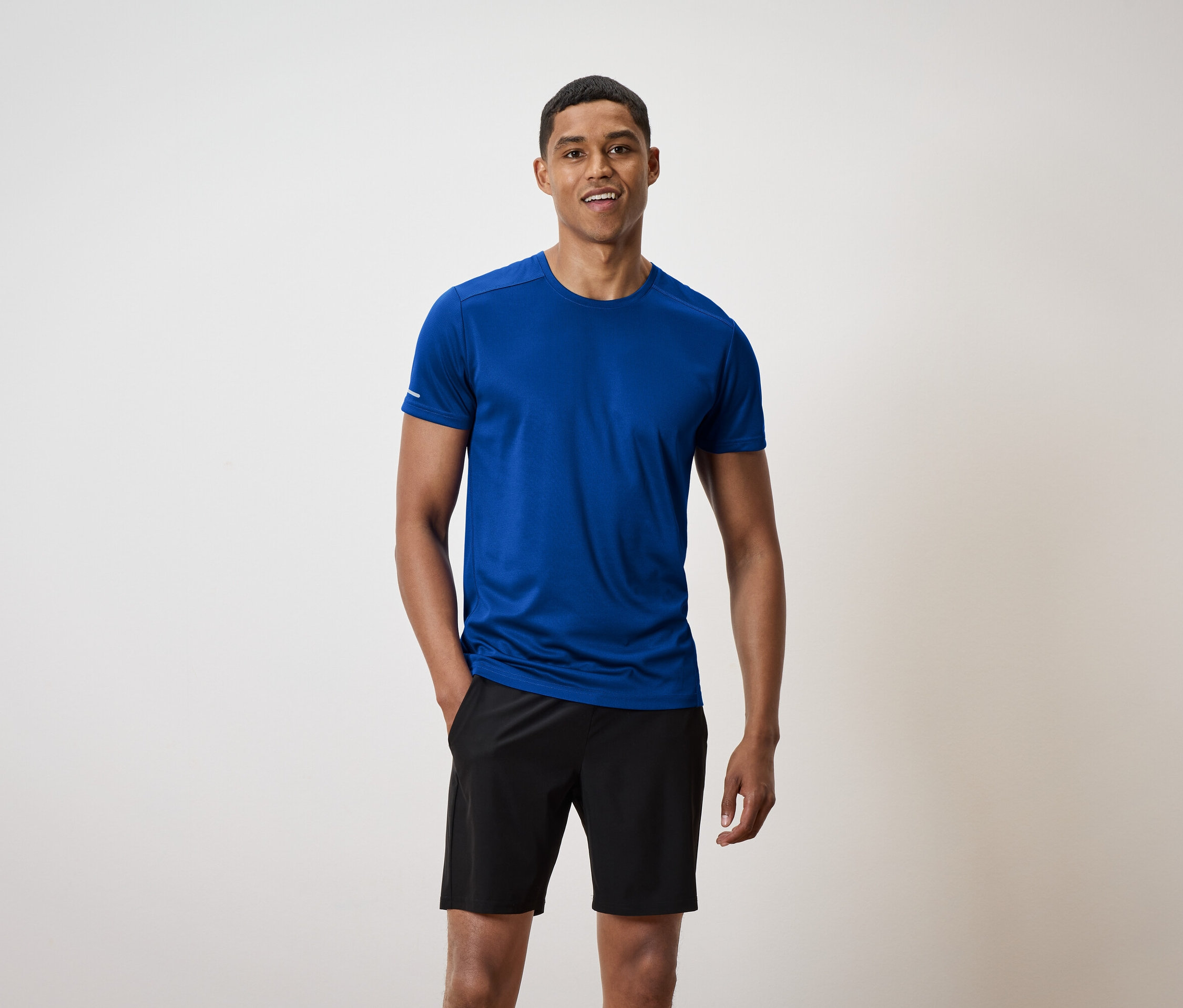 Ein kurzhaariger Mann steht und lächelt, er trägt ein blaues T-Shirt und schwarze Shorts.