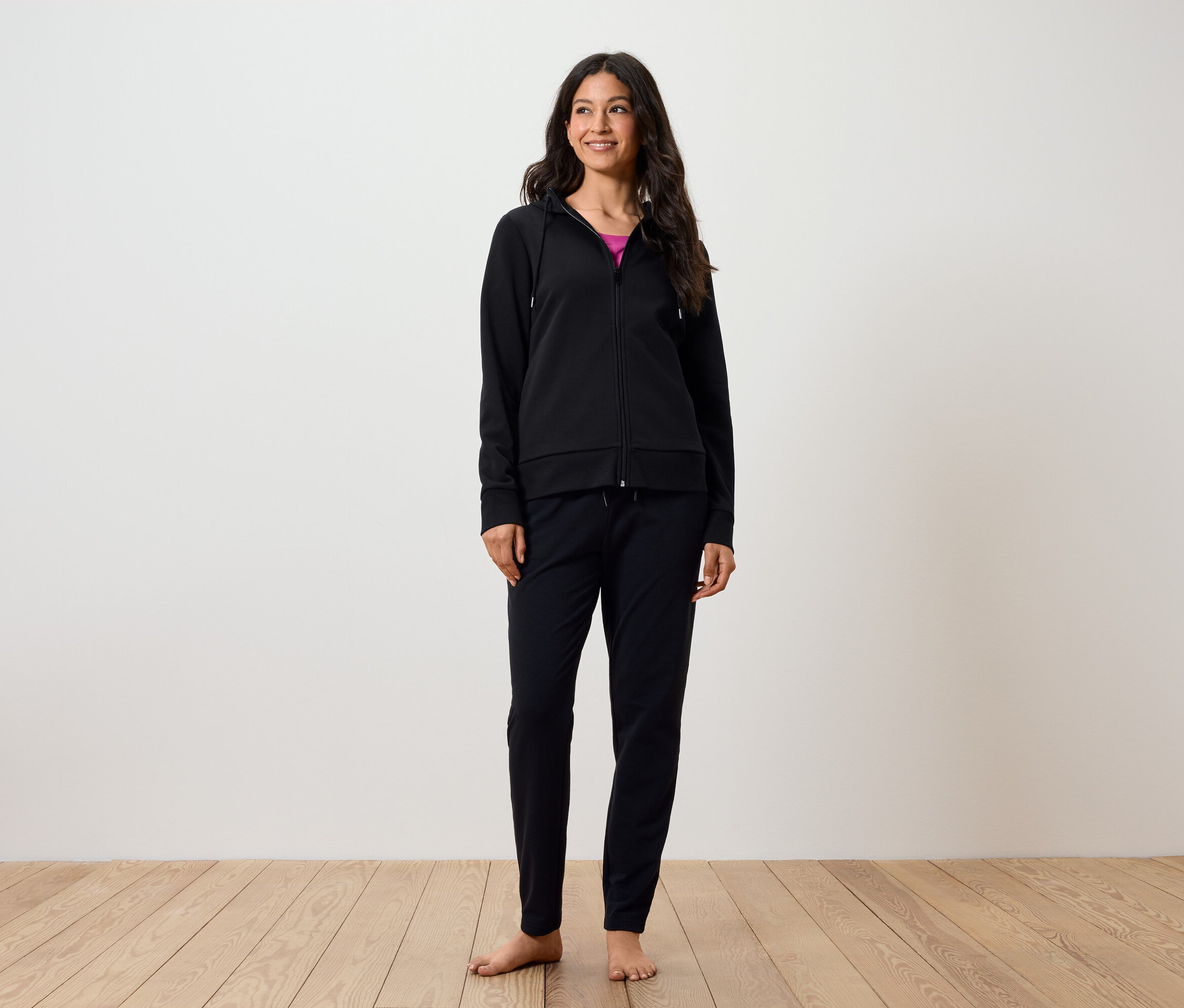 Eine lächelnde Frau steht barfuß auf einem Holzboden. Sie trägt eine schwarze Hose und einen schwarzen Kapuzenpullover mit Reißverschluss.