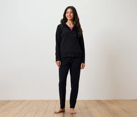 Eine lächelnde Frau steht barfuß auf einem Holzboden. Sie trägt eine schwarze Hose und einen schwarzen Kapuzenpullover mit Reißverschluss.