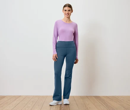 Frau in lila Langarmshirt und blauen Hosen steht auf Holzboden vor einer weißen Wand.