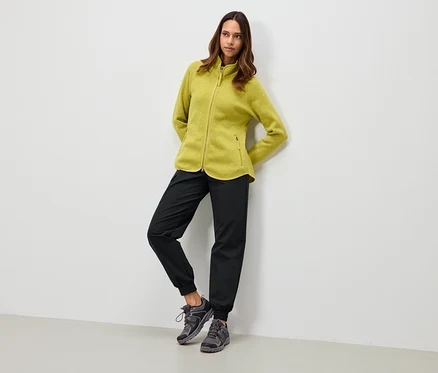 Frau in olivgrüner Fleecejacke, schwarzer Hose und grauen Schuhen, die sich an eine weiße Wand lehnt.