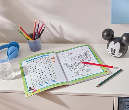 Auf einem weißen Tisch liegen ein aufgeschlagenes Malbuch, bunte Stifte in einem blauen Becher, blaue Kopfhörer, ein Glas Wasser und eine Mickey Mouse-Figur.