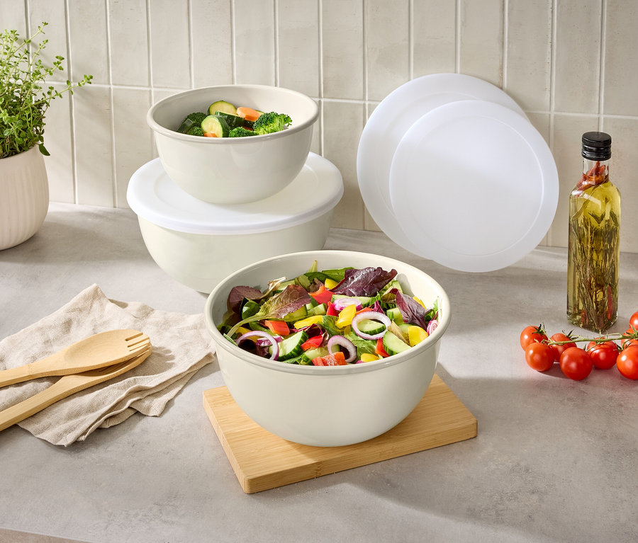 Auf einer Küchentheke stehen Schüsseln mit Salat und Gemüse, daneben ein Kräutertopf, Salatbesteck, Tomaten, Öl und Deckel.