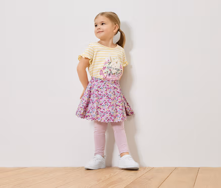 Ein kleines Mädchen mit blonden Haaren trägt ein gelb-gestreiftes Shirt mit einem Blumenmuster, einen geblümten Rock, rosa Leggings und weiße Turnschuhe.