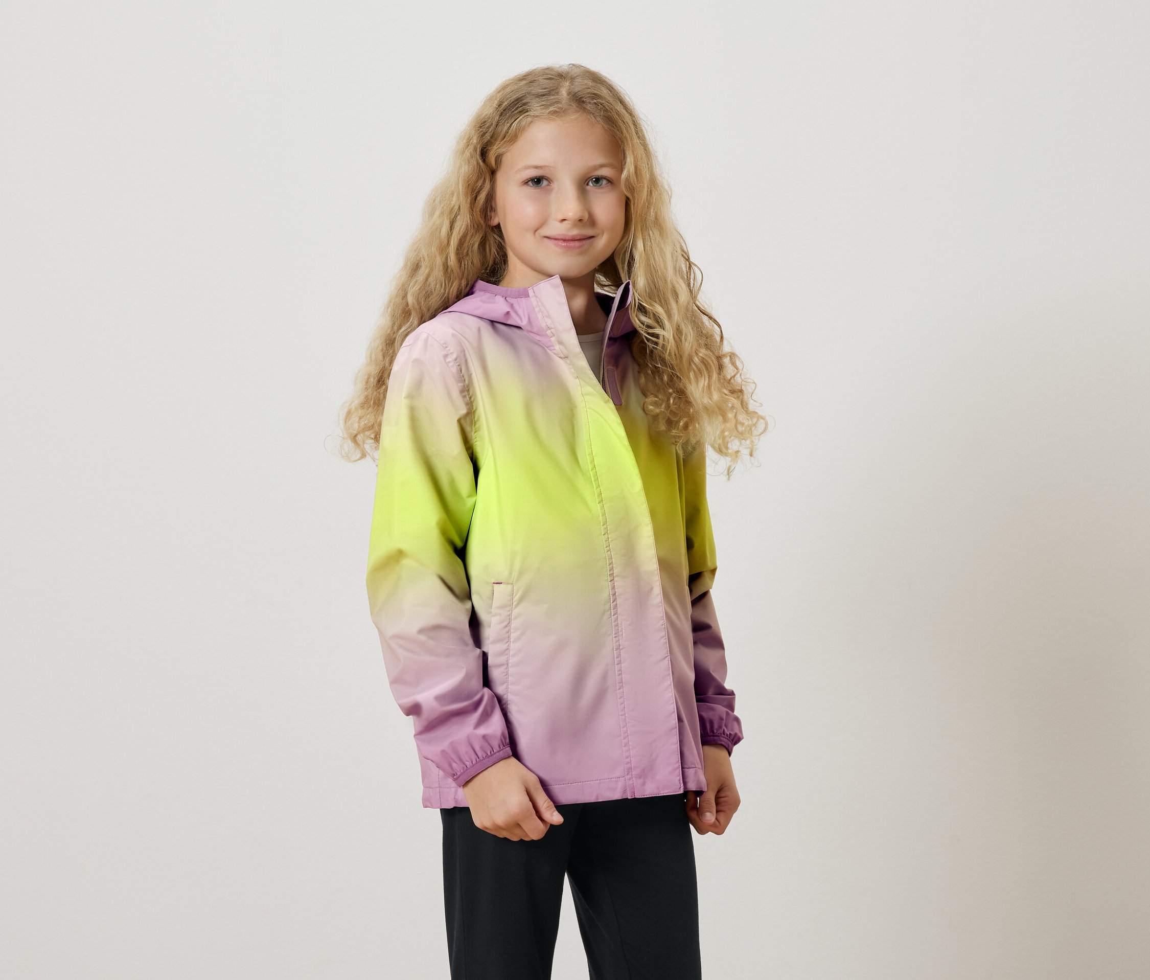 Mädchen mit langen blonden Locken trägt eine Kapuzenjacke mit Farbverlauf von Gelb nach Lila.