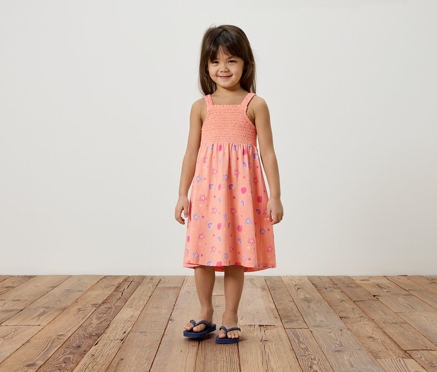 Mädchen mit braunen Haaren trägt ein rosa Sommerkleid mit Sternen und Herzchen und blaue Flip-Flops.