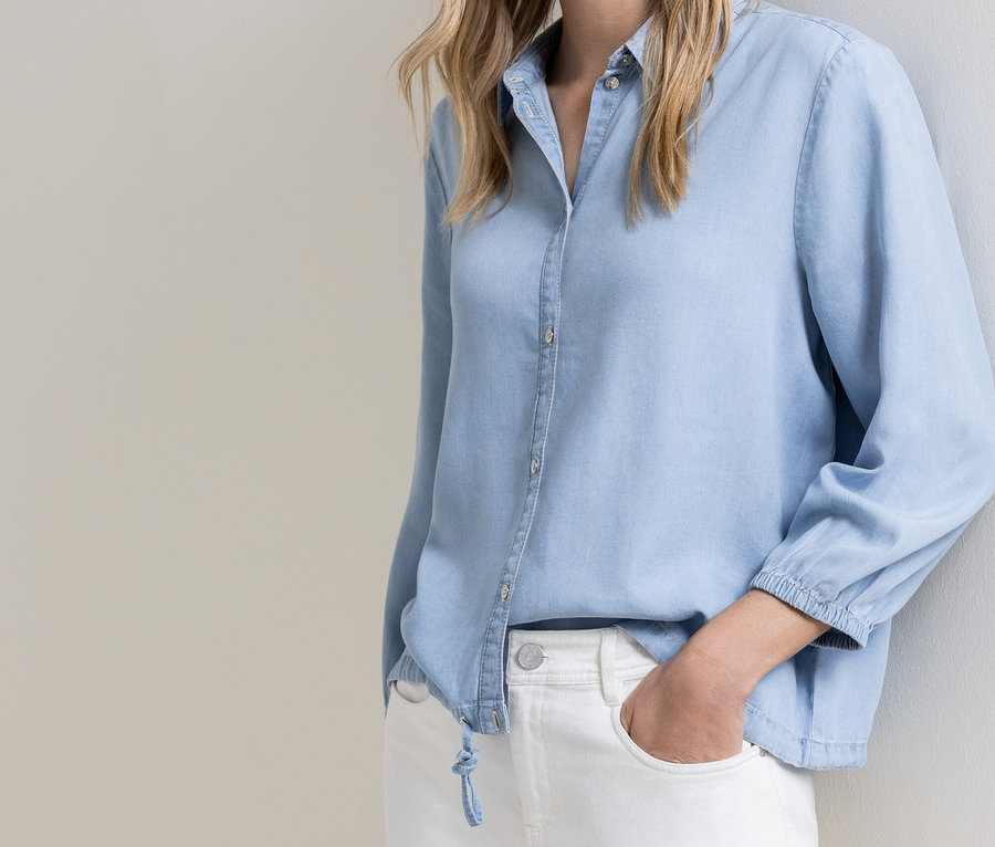 Frau mit hellblauer Knopfleiste und weißen Jeans, eine Hand in der Hosentasche, an einer Wand lehnend.