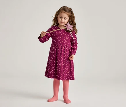 Ein Mädchen mit lockigem Haar hält einen Zauberstab und trägt ein burgunderrotes Kleid mit Blumenmuster und rosa Strumpfhosen.