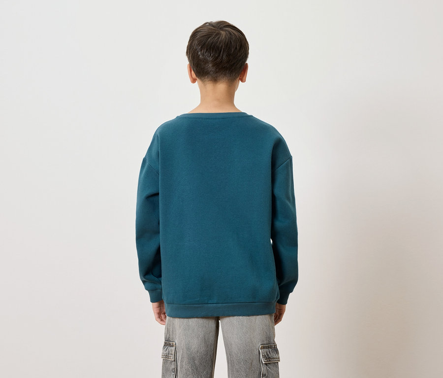 Rückansicht eines Kindes mit blaugrünem Pullover und grauen Shorts vor einem weißen Hintergrund.