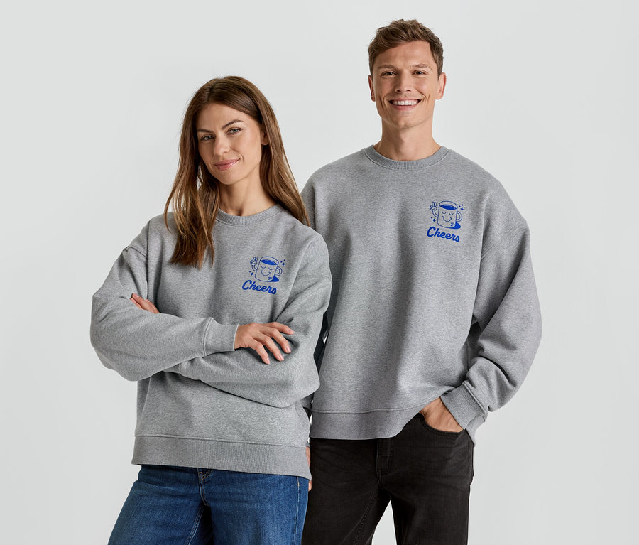 Eine Frau mit verschränkten Armen und ein Mann stehen nebeneinander und tragen graue Pullover mit einem blauen "Cheers"-Aufdruck.