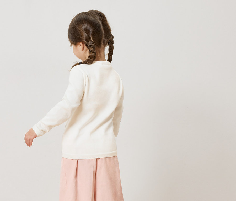 Rückansicht eines kleinen Mädchens mit Zöpfen, das einen weißen Pullover und einen rosa Rock trägt.