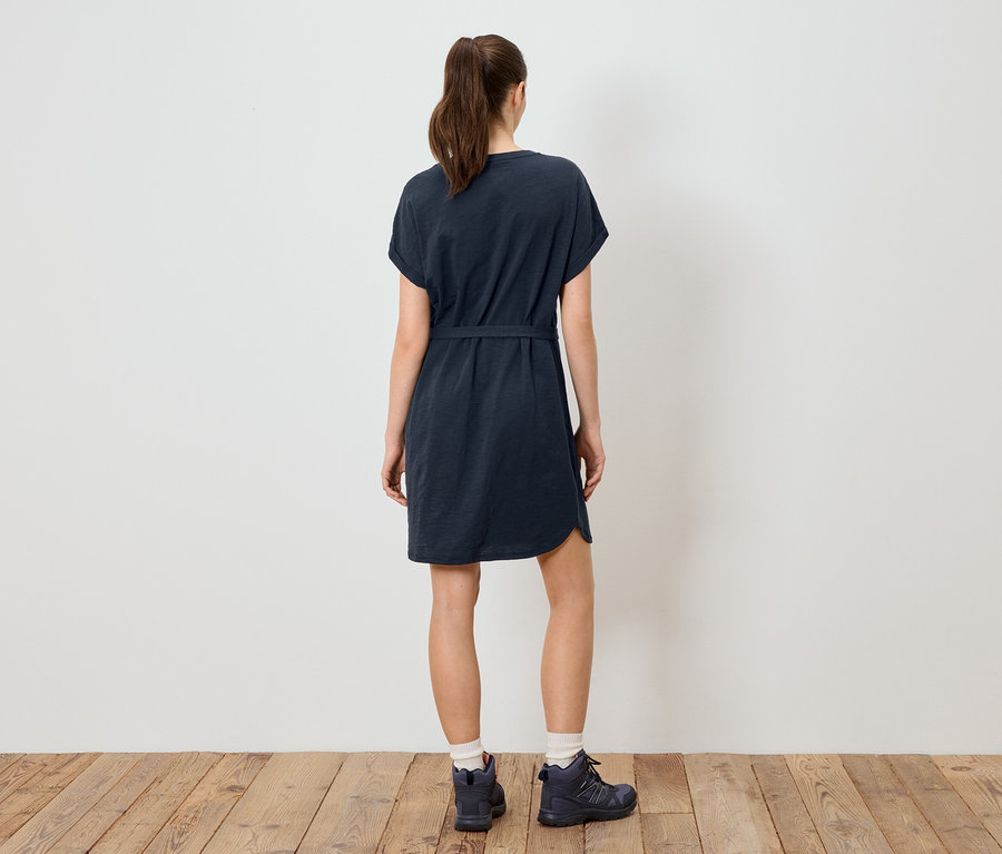 Rückansicht einer Frau mit Pferdeschwanz, die ein marineblaues T-Shirt-Kleid mit Gürtel und Wanderschuhe vor einer weißen Wand auf einem Holzboden trägt.
