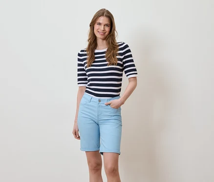 Eine Frau steht vor einem hellen Hintergrund, sie trägt ein gestreiftes T-Shirt und hellblaue Shorts.