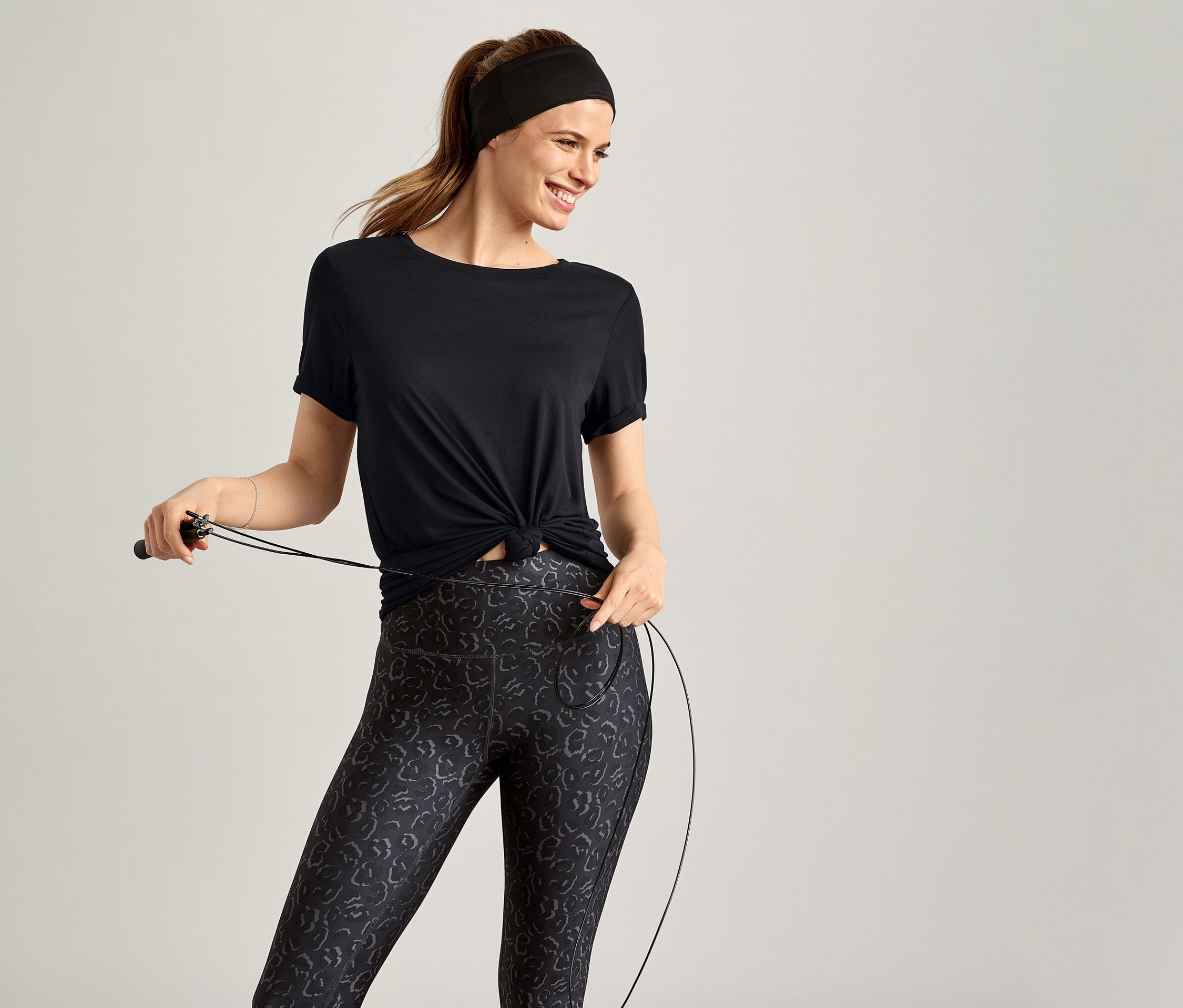 Frau mit Stirnband und Sportshirt, schwarz, hält ein Springseil vor grauem Hintergrund.