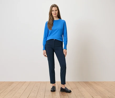 Frau in blauer Bluse und marineblauer Hose, dazu schwarze Slipper. Sie steht auf einem Holzboden vor einer weißen Wand.