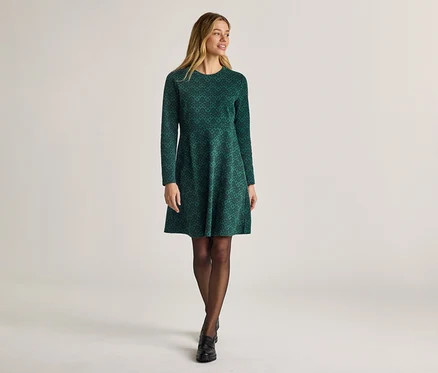 Frau in einem grünen, gemusterten, langärmeligen Kleid, Strumpfhose und Schuhen vor einem hellen Hintergrund.