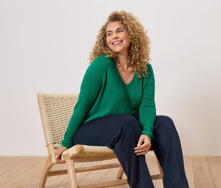 Frau mit lockigen Haaren sitzt auf einem Stuhl und trägt einen grünen Pullover und eine blaue Hose.