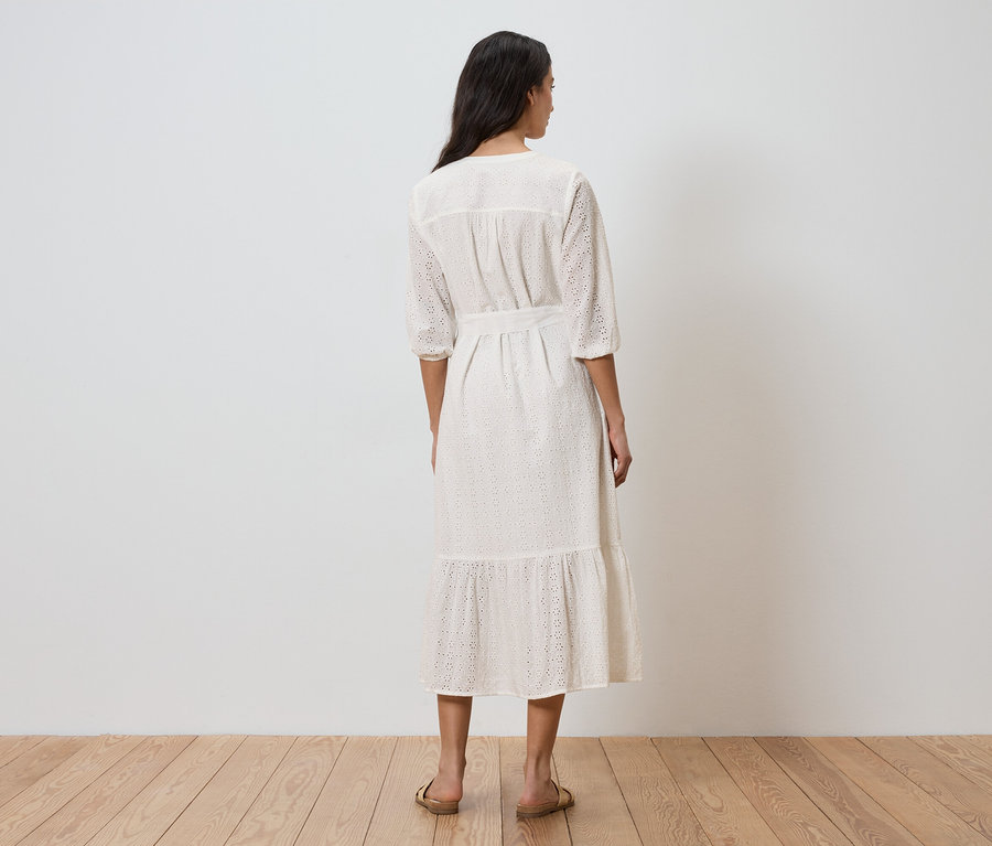 Eine Frau steht mit dem Rücken zur Kamera und trägt ein weißes Kleid mit Gürtel und Lochmuster.