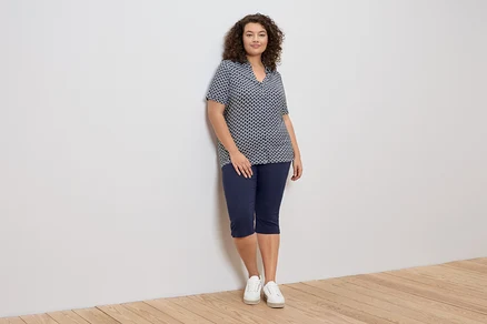 Eine Frau mit lockigem Haar lehnt an einer Wand und trägt eine blau-weiß gemusterte Bluse, blaue Caprihosen und weiße Turnschuhe.