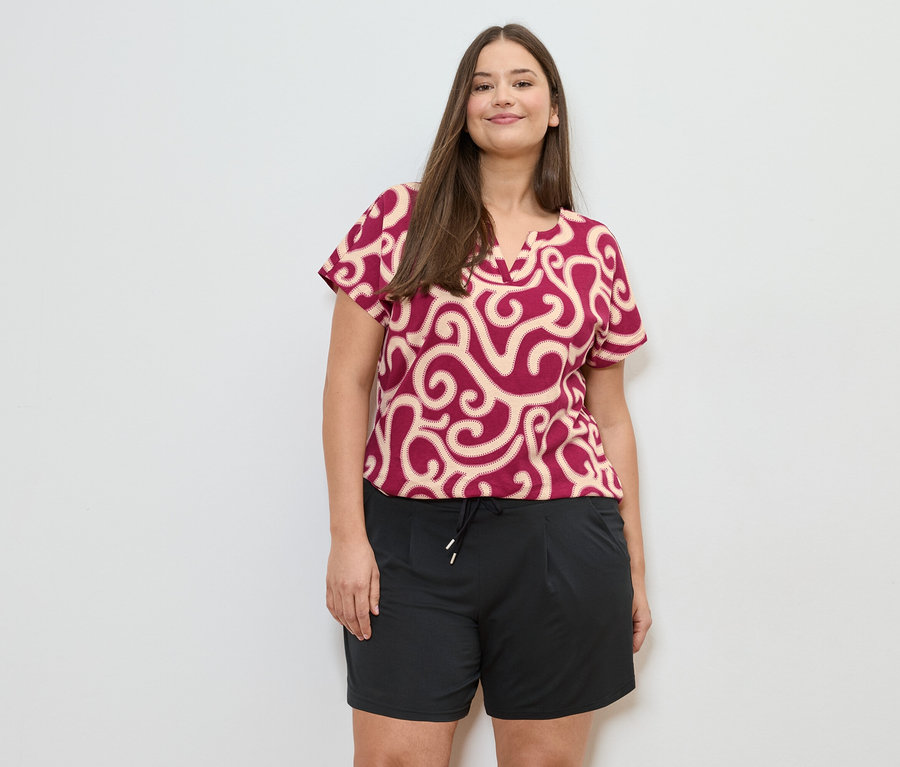 Eine Frau mit braunen Haaren trägt eine rot-beige gemusterte Bluse und schwarze Shorts vor einem weißen Hintergrund.