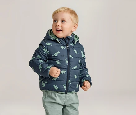 Kleiner Junge mit blonden Haaren trägt eine marineblaue, gesteppte Jacke mit Dinosaurier-Muster und grüne Hosen.