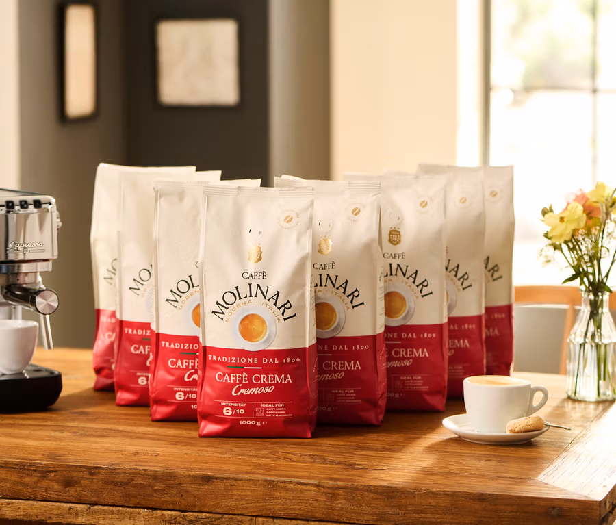 Auf einem Holztisch stehen eine Espressomaschine neben einer Tasse mit Kaffee und einem Keks. Dahinter befinden sich acht Packungen Caffè Molinari Caffè Crema Cremoso - 8x 1 kg Ganze Bohne und eine Vase mit Blumen.