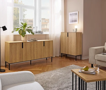 Ein Wohnzimmer mit einem langen Sideboard aus hellem Holz unter einem Fenster und einem passenden hohen Schrank. Eine schwarze Stehlampe, zwei Sessel, ein zotteliger Teppich und ein Couchtisch mit einer Birne und einem Wasserglas sind ebenfalls zu sehen.