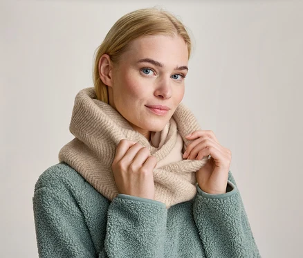 Blonde Frau mit blauen Augen trägt einen beigefarbenen Strick-Rundschal und eine mintgrüne Fleecejacke, hält den Schal mit beiden Händen und blickt nach vorne.