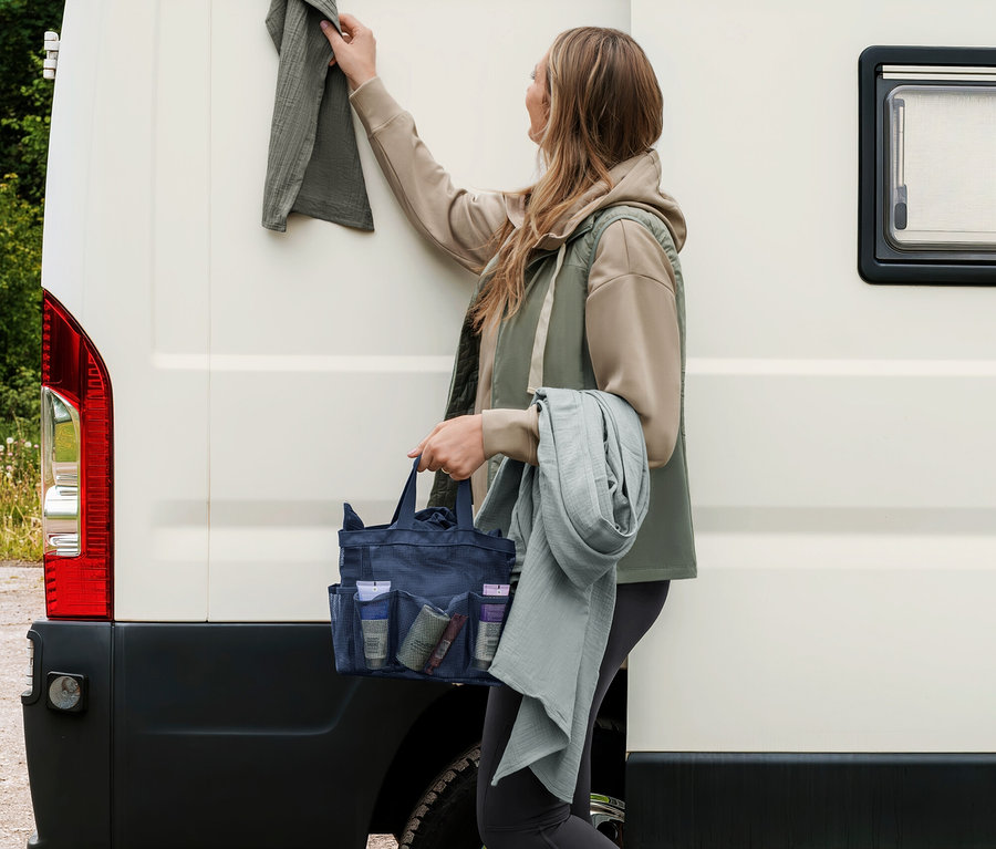 Frau hängt ein graugrünes Musselin-Handtuch an einen Campervan, hält eine dunkelblaue Duschtasche und ein weiteres hellgraugrünes Musselin-Handtuch.